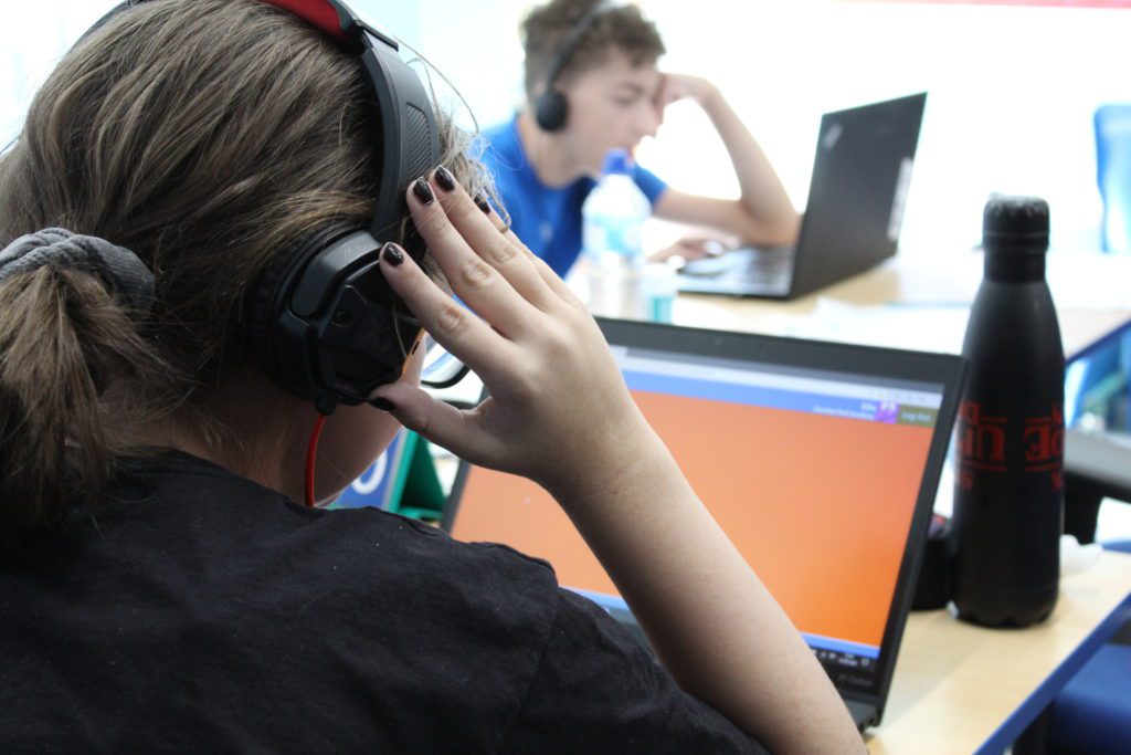 girl at deskwith headphones and laptop with boy in the distance on laptop