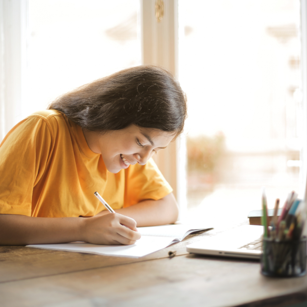 Girl in yellow top at desk writing next to a laptop