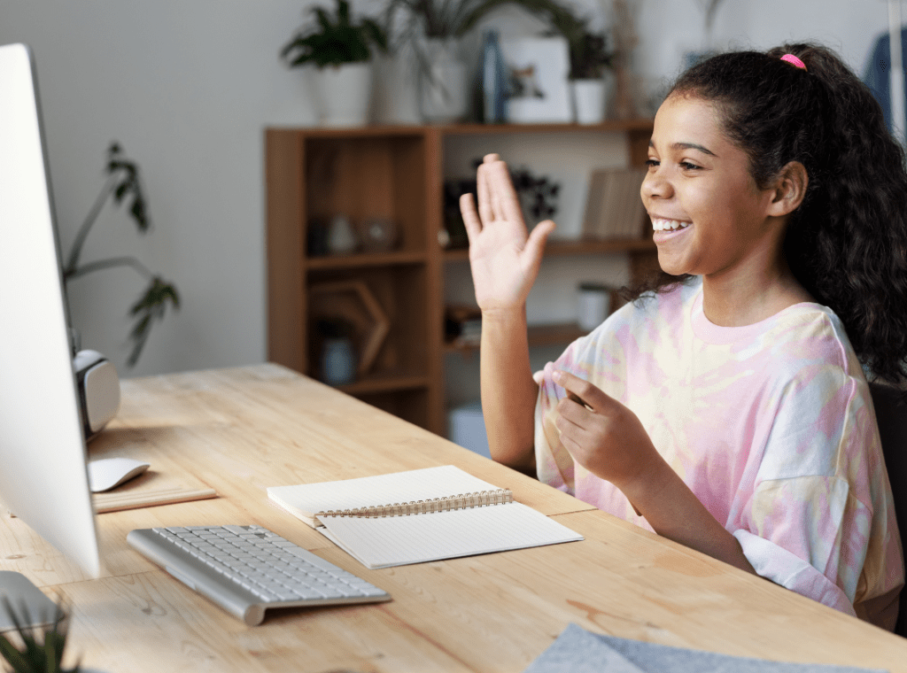 Child smiling looking at a computer screen, raising her hand at her desk